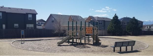 An image of a playground that has a slide, pole, stairs, and tick-tack-toe blocks. The playground is green and beige and has a nearby bench and surrounded by grass.